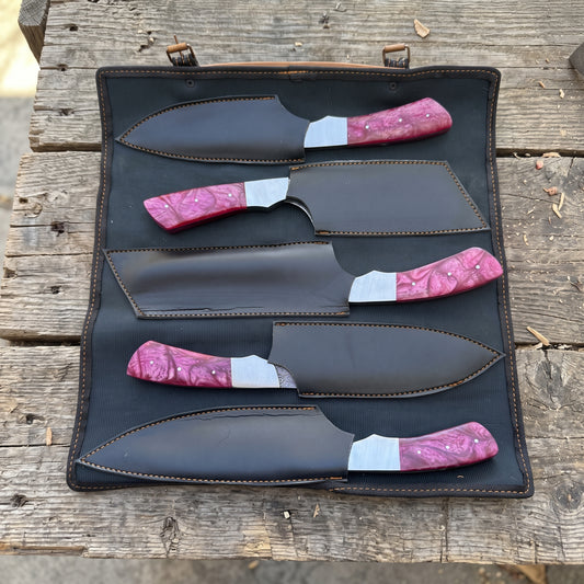 Five fixed-blade chef knives with red resin handles and black sheaths arranged in a black leather roll-up case on a rustic wooden surface.