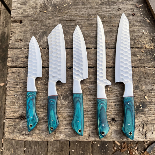 Five kitchen chef knives with hammered white steel blades and turquoise resin handles, arranged in descending size order on a rustic wooden surface.
