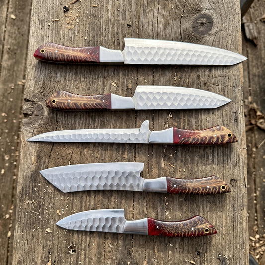 Five fixed-blade knives with hammered steel blades and reddish-brown patterned handles are arranged vertically on a rustic wooden plank outdoors.