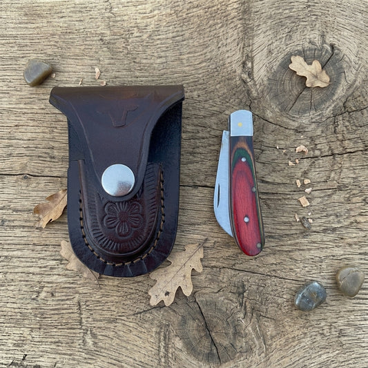 A leather sheath and folding pocket knife displayed on a rustic wood surface with scattered oak leaves and small stones. The sheath is dark brown leather with an embossed floral pattern and a silver snap closure.