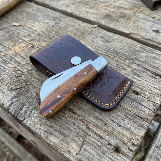 A folding pocket knife with a wood-grain handle and silver blade, resting on a dark brown tooled leather sheath over a rustic wooden plank surface.