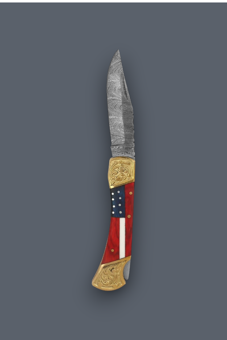 A custom folding pocket knife with a Damascus steel blade, gold-toned brass bolsters, and a handle featuring a red, white, and blue American flag design with brass pins, shown on a solid dark gray background.
