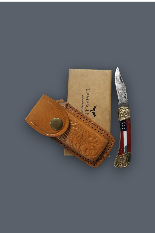 A folding pocket knife with a red, white, and blue striped handle and ornate gold-toned end pieces, displayed next to its tan leather sheath and a tan paper box printed with the Damace logo, all on a solid dark gray background.
