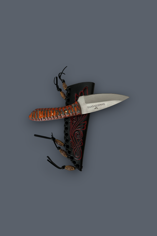 A fixed-blade knife with a silver blade and a textured orange-brown handle, displayed alongside a black sheath with red tribal-style patterns and black cords, set against a solid dark gray background.