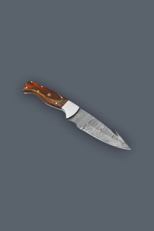 Angled flat lay of a Damascus steel fixed blade knife featuring a spine gut hook, polished metal bolster, and a custom wood handle.