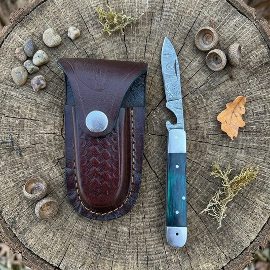 A folding pocket knife with a patterned steel blade and a blue-green handle is displayed next to its dark brown leather sheath on a rustic tree stump, surrounded by small stones, acorns, and dried leaves.