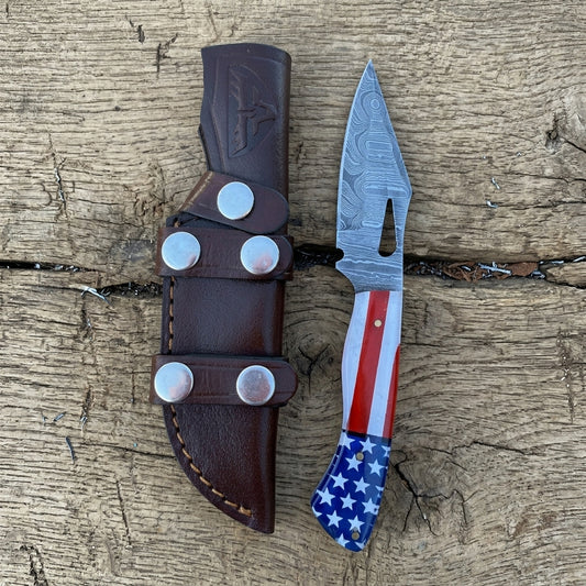 A custom fixed blade knife with a patterned steel blade and an American flag handle, displayed next to its brown leather sheath on a rustic wooden surface.