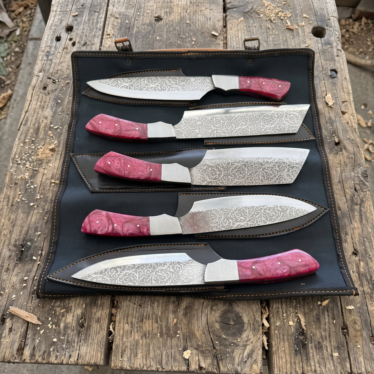 Four patterned steel chef knives set with red resin handles are displayed in a black leather roll-up case on a rustic wooden surface.