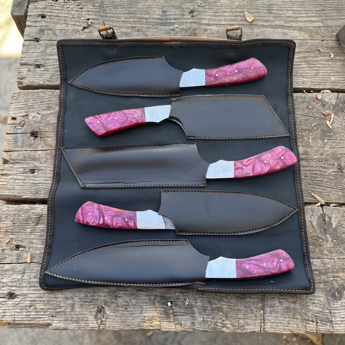 Five fixed-blade chef knives with red resin handles and black sheaths arranged in a black leather roll-up case on a rustic wooden surface.