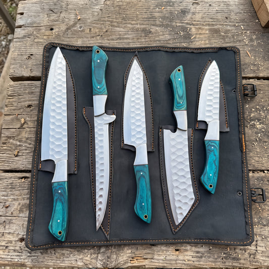 Five fixed-blade chef knives with hammered silver blades and turquoise-colored handles are displayed in a black leather knife roll on a rustic wooden surface.