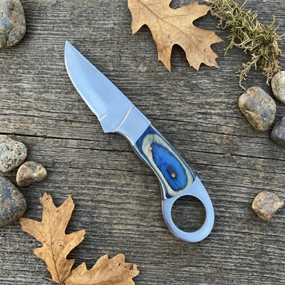 A fixed-blade knife with a satin silver blade and a blue marbled handle, displayed diagonally on a rustic weathered wood surface surrounded by autumn leaves, moss, and small stones.