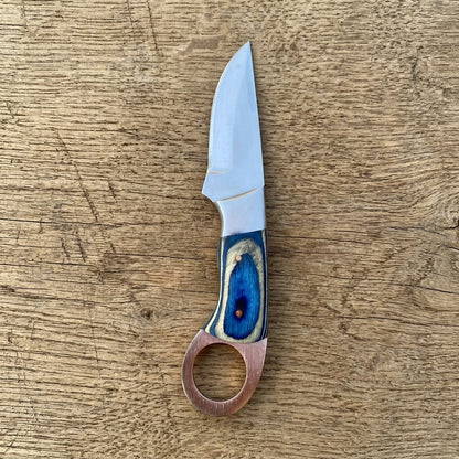 A small fixed-blade knife with a silver steel blade and a decorative handle made of layered blue, beige, and copper-colored wood, placed on a rustic wooden surface.