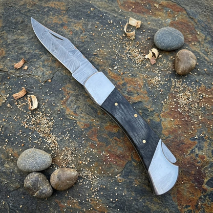 A folding pocket knife with a Damascus steel blade and a black handle with brass pins, displayed diagonally on a textured stone surface with small wood shavings and stones.