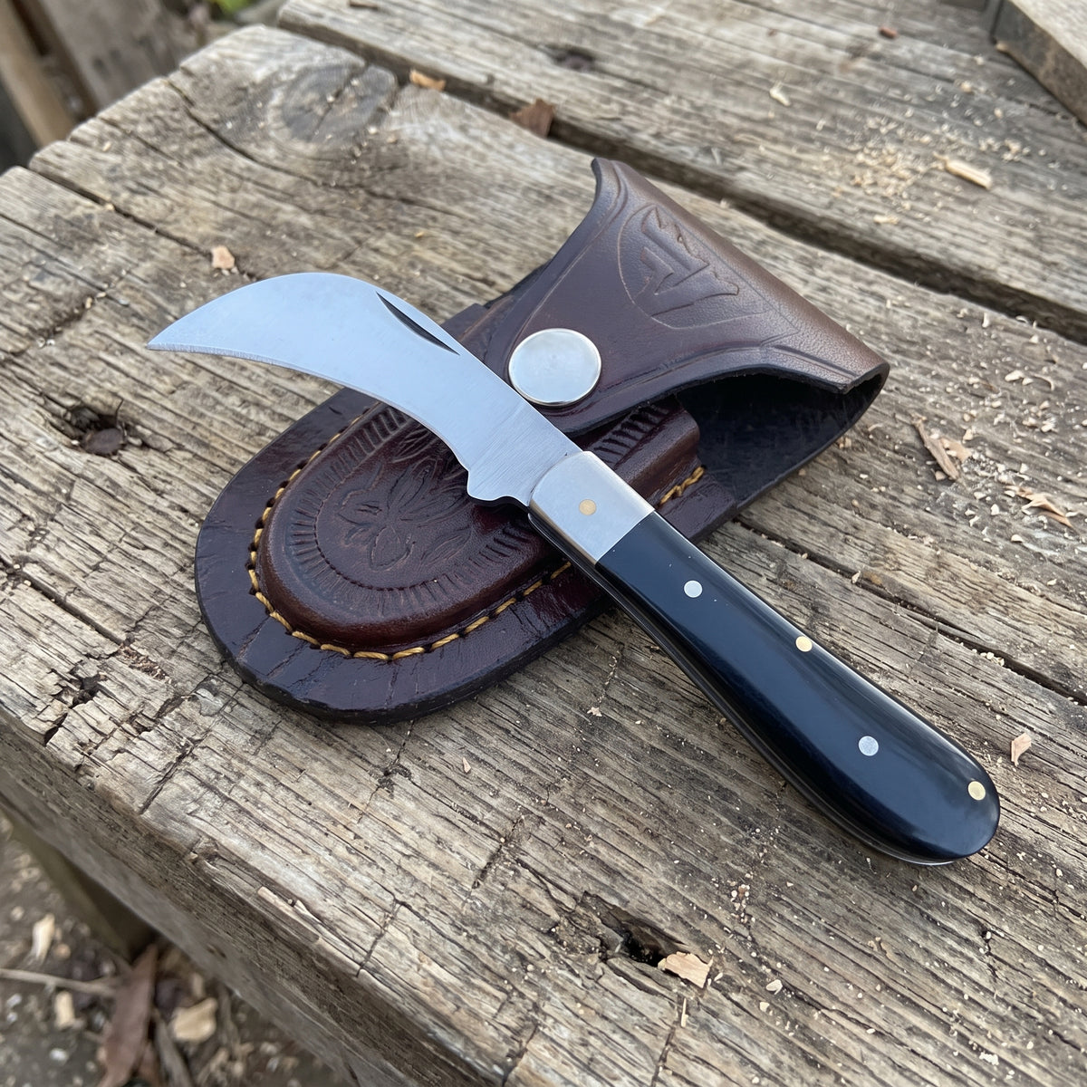 A folding pocket knife with a curved silver blade and black handle, resting on a brown leather sheath over a weathered wooden surface.