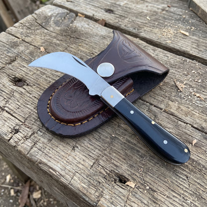 A folding pocket knife with a curved silver blade and black handle, resting on a brown leather sheath over a weathered wooden surface.