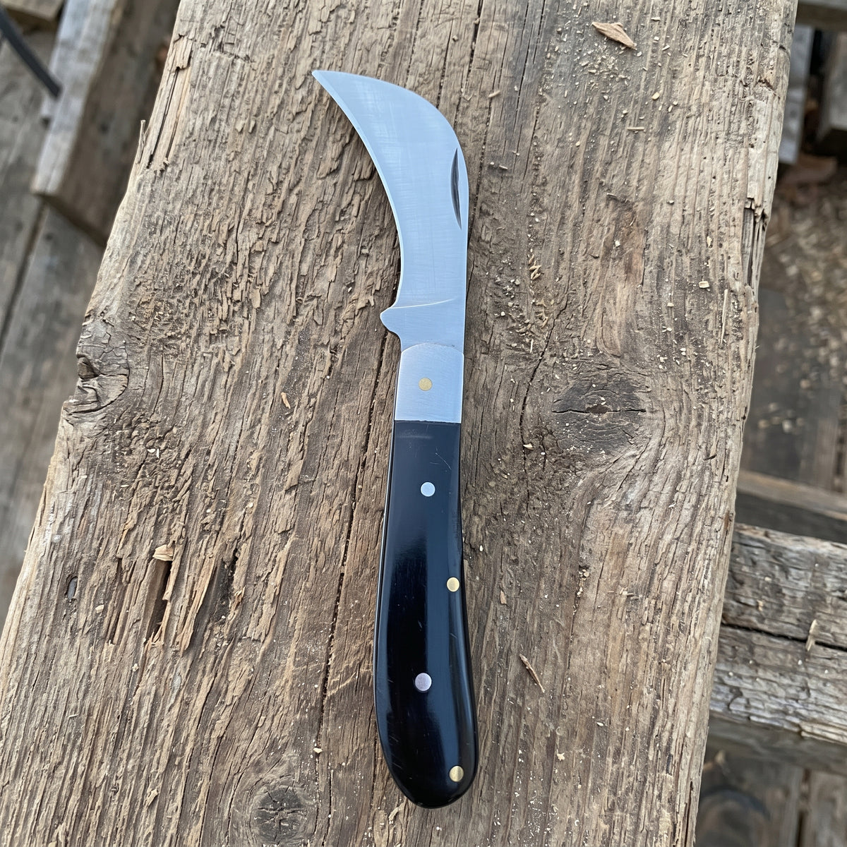 A curved-handled knife with a silver steel blade and a black handle secured by brass rivets, resting on a weathered wooden plank.