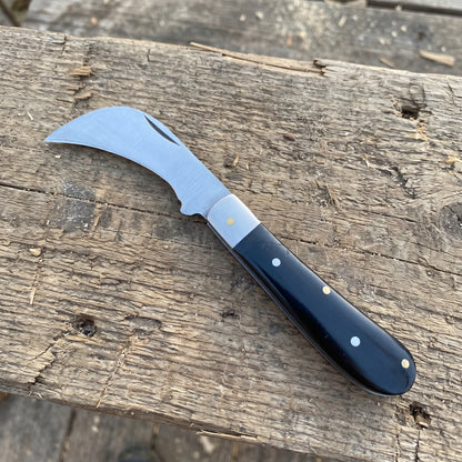 A curved pocket knife with a silver steel blade and a dark navy blue handle with brass and white dots, resting on a rustic weathered wooden surface.