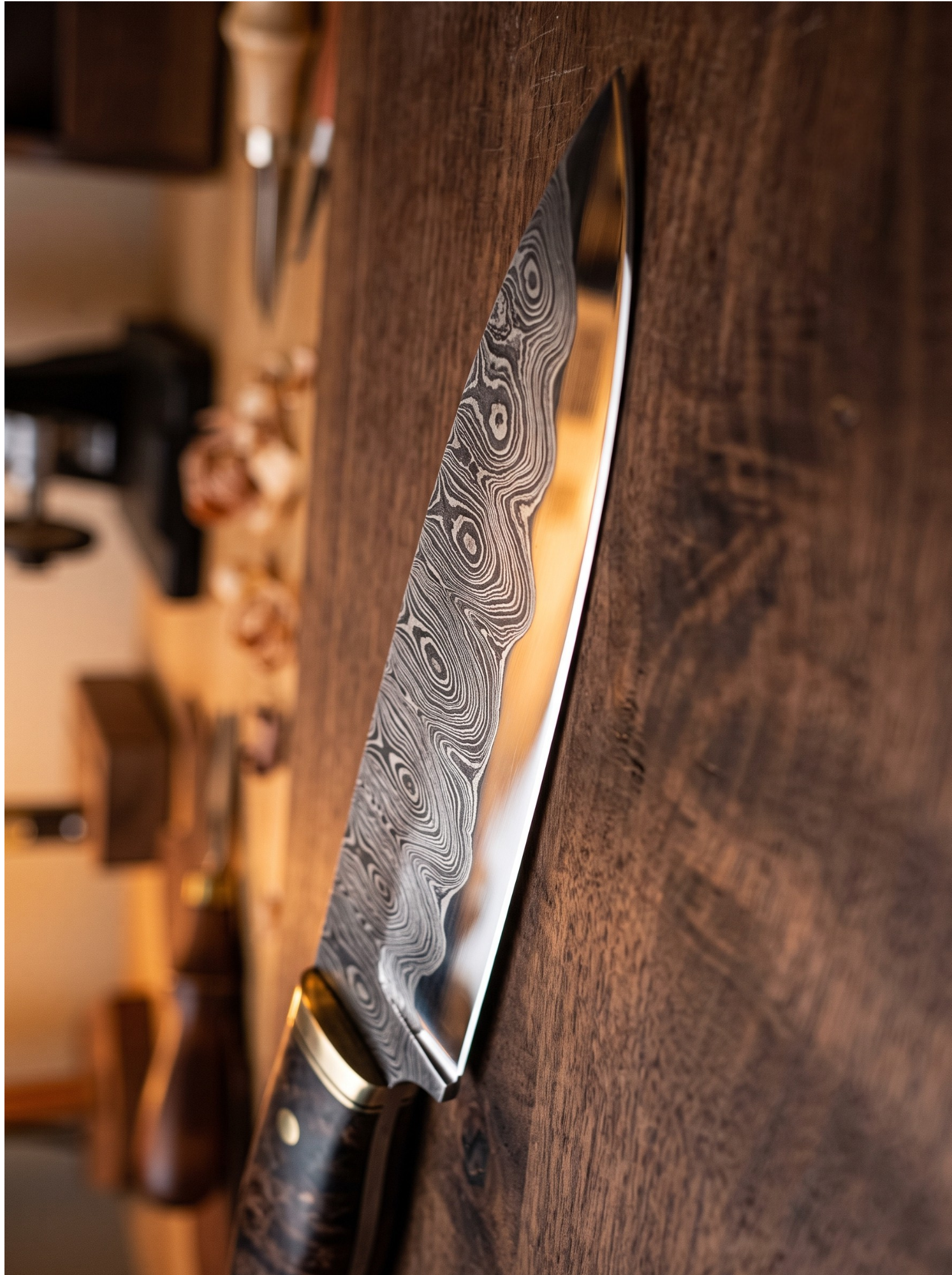 A Damascus steel chef's knife with a patterned steel blade and a dark wood handle with brass bolsters, resting on a rustic dark wood table with a blurred workshop background.