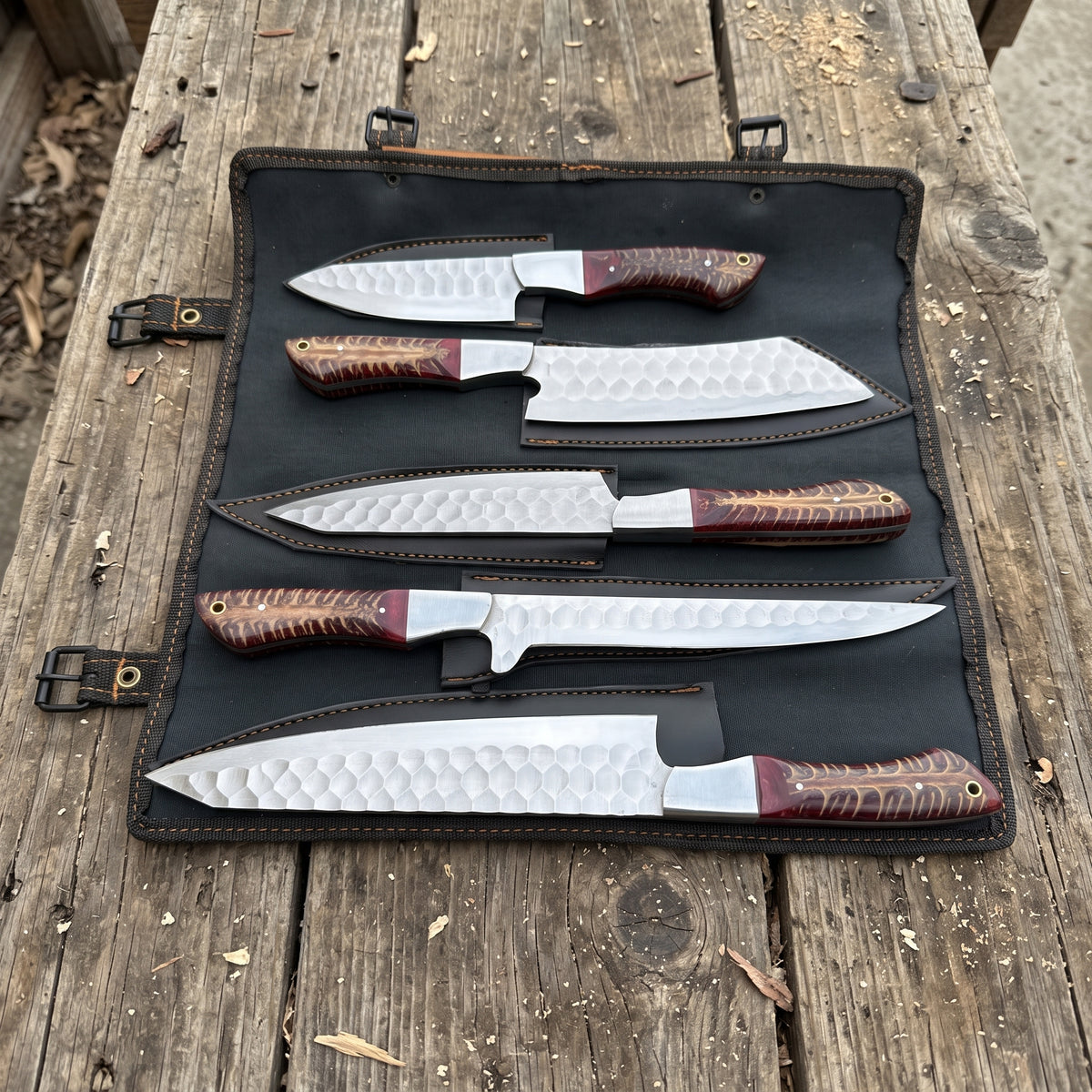 Five kitchen knives with patterned steel blades and reddish-brown handles are displayed in an open black leather roll-up case on a rustic wooden table.