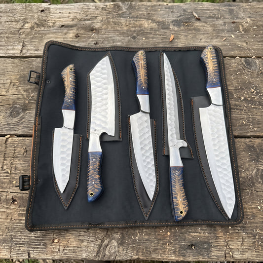 Five kitchen knives with hammered silver blades and blue-and-gold patterned handles, displayed in a black roll-up knife case on a rustic wooden surface.