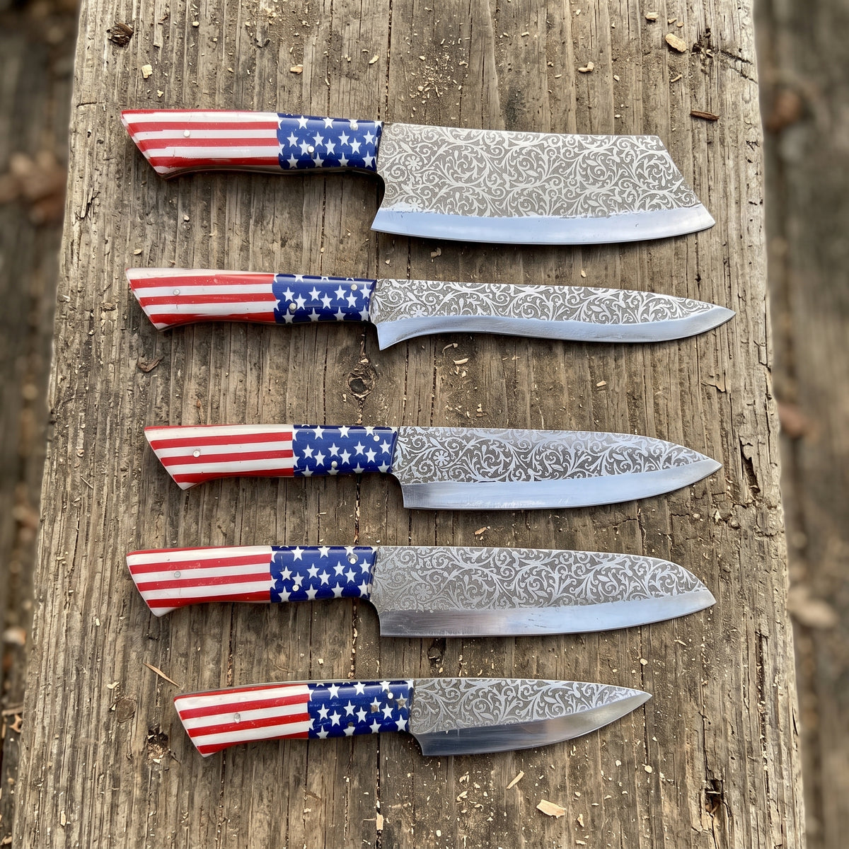 Five kitchen knives with patterned steel blades and American flag handles are arranged vertically on a rustic wooden plank outdoors.