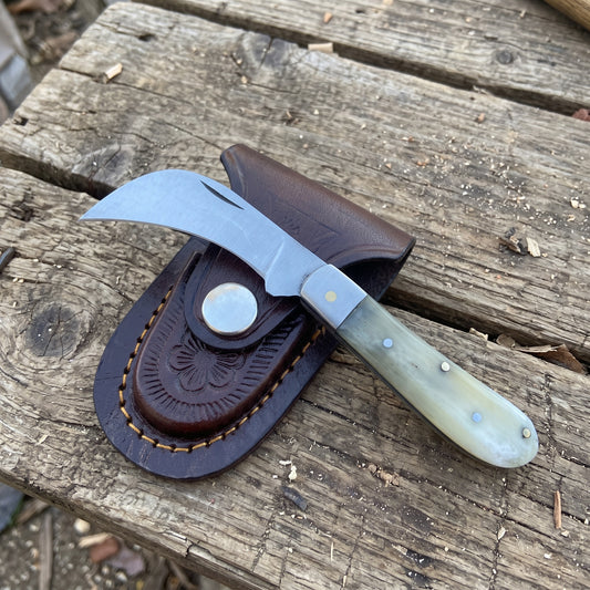 A folding pocket knife with a curved silver blade and a pale green bone handle, resting on a dark brown leather sheath over a rustic wooden plank background.