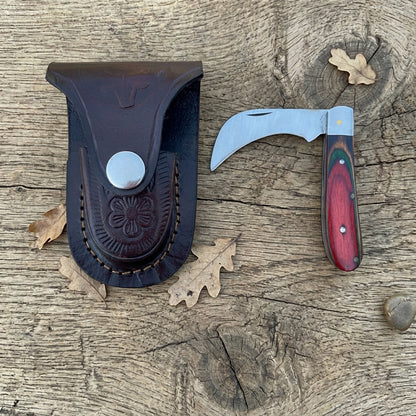 A folding pocket knife with a curved silver blade and reddish-brown wood handle, displayed next to its dark brown leather sheath on a rustic wooden surface with scattered oak leaves.