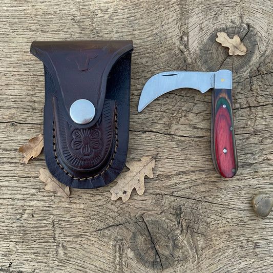 A folding pocket knife with a curved silver blade and reddish-brown wood handle, displayed next to its dark brown leather sheath on a rustic wooden surface with scattered oak leaves.