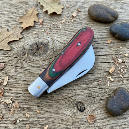 A folding pocket knife with a red and green wood handle and a silver metal blade, resting on a rustic weathered wood surface with scattered leaves, wood shavings, and stones.
