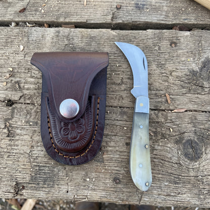A folding pocket knife with a curved silver blade and a cream-colored bone handle, displayed next to its dark brown leather sheath on a rustic wooden surface.