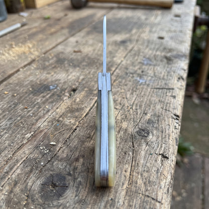 A folding pocket knife with a polished silver blade and a marbled bone handle in beige, brown, and gray tones, standing upright on a rustic wooden workbench.
