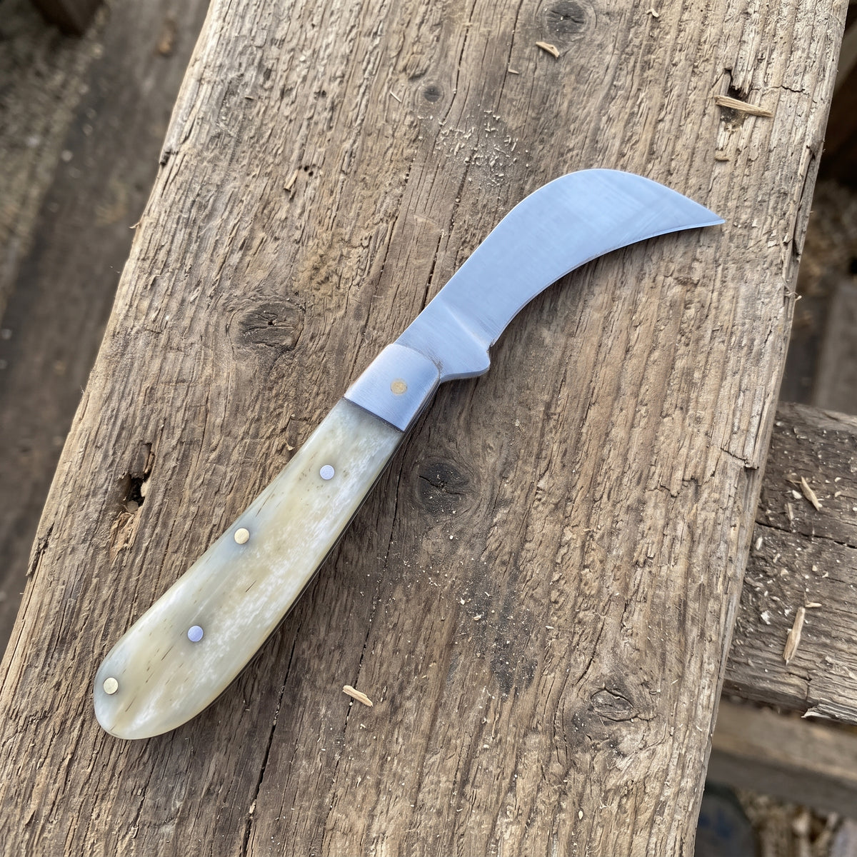 A curved knife with a silver steel blade and a cream-colored bone handle, resting diagonally on a rustic wooden surface.