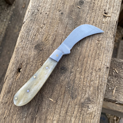 A curved knife with a silver steel blade and a cream-colored bone handle, resting diagonally on a rustic wooden surface.