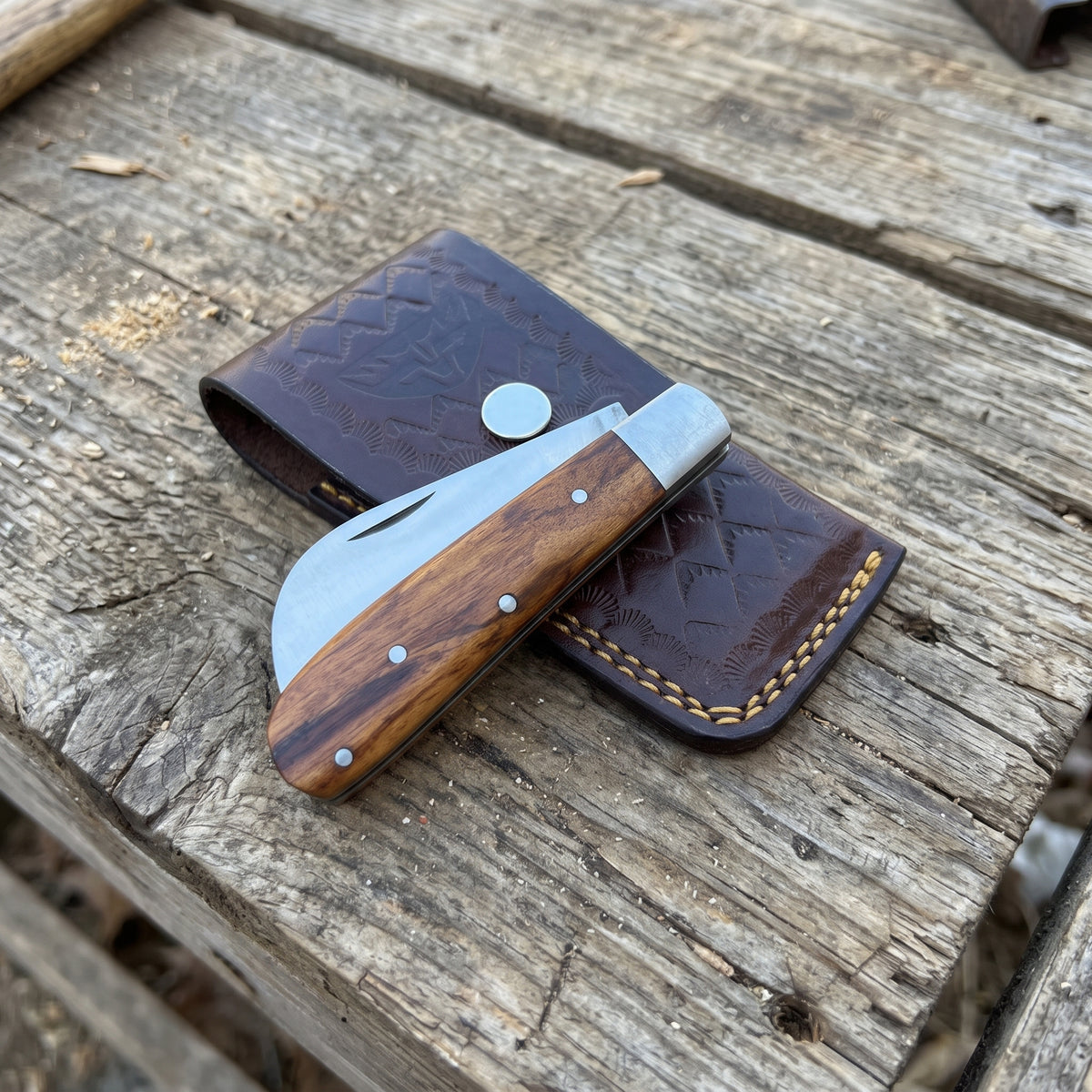 A folding pocket knife with a wood-grain handle and silver blade, resting on a dark brown tooled leather sheath over a rustic wooden plank surface.