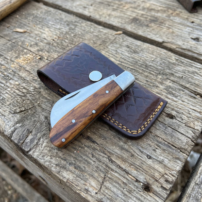 A folding pocket knife with a wood-grain handle and silver blade, resting on a dark brown tooled leather sheath over a rustic wooden plank surface.