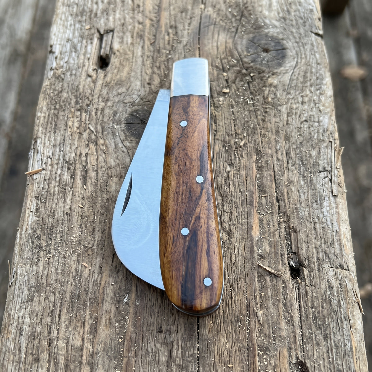 A folding pocket knife with a dark brown wood handle and silver metal blade, resting on a rustic weathered wooden log.