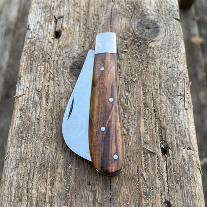A folding pocket knife with a dark brown wood handle and silver metal blade, resting on a rustic weathered wooden log.