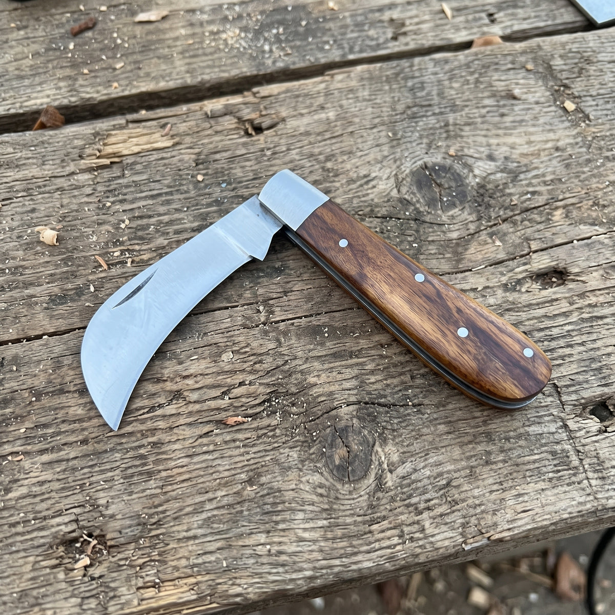 A folding pocket knife with a curved silver steel blade and a brown wooden handle secured by silver rivets, resting on a weathered wooden plank surface.