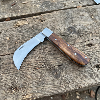 A folding pocket knife with a curved silver steel blade and a brown wooden handle secured by silver rivets, resting on a weathered wooden plank surface.