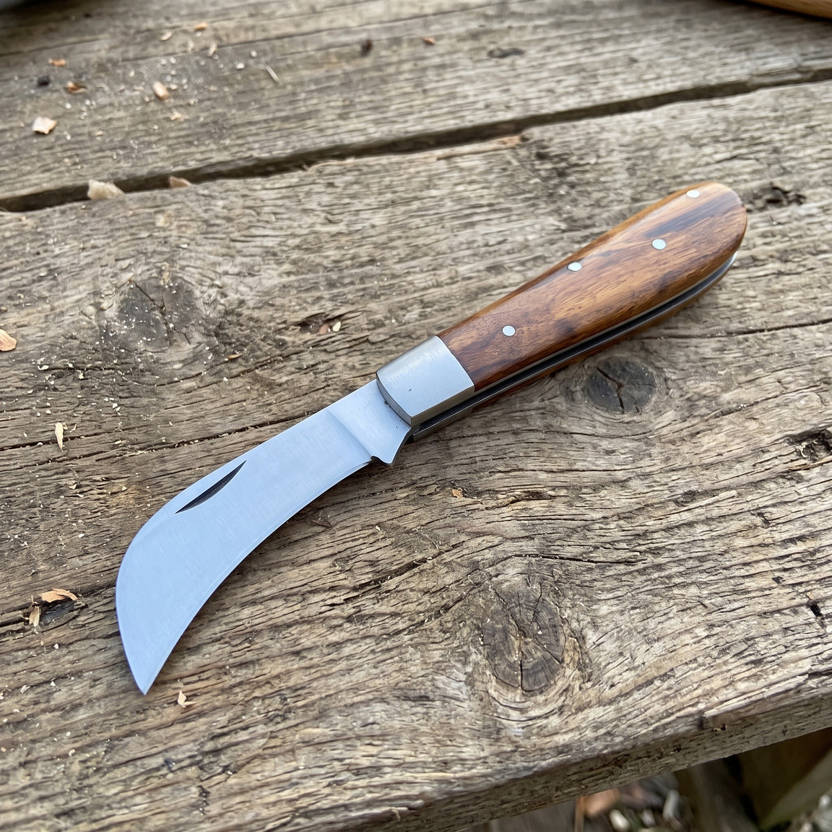 A pocket knife with a curved silver steel blade and a brown wooden handle, resting on a rustic weathered wooden plank surface.
