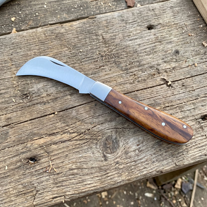A curved-handled knife with a silver steel blade and a polished brown wooden handle, resting diagonally on a rustic weathered wooden plank surface.