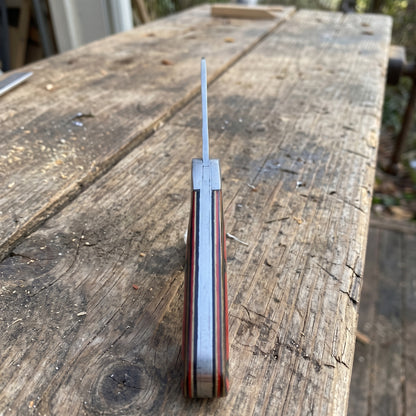 A folding pocket knife with a striped red, brown, and silver handle, shown partially open on a rustic wooden workbench.