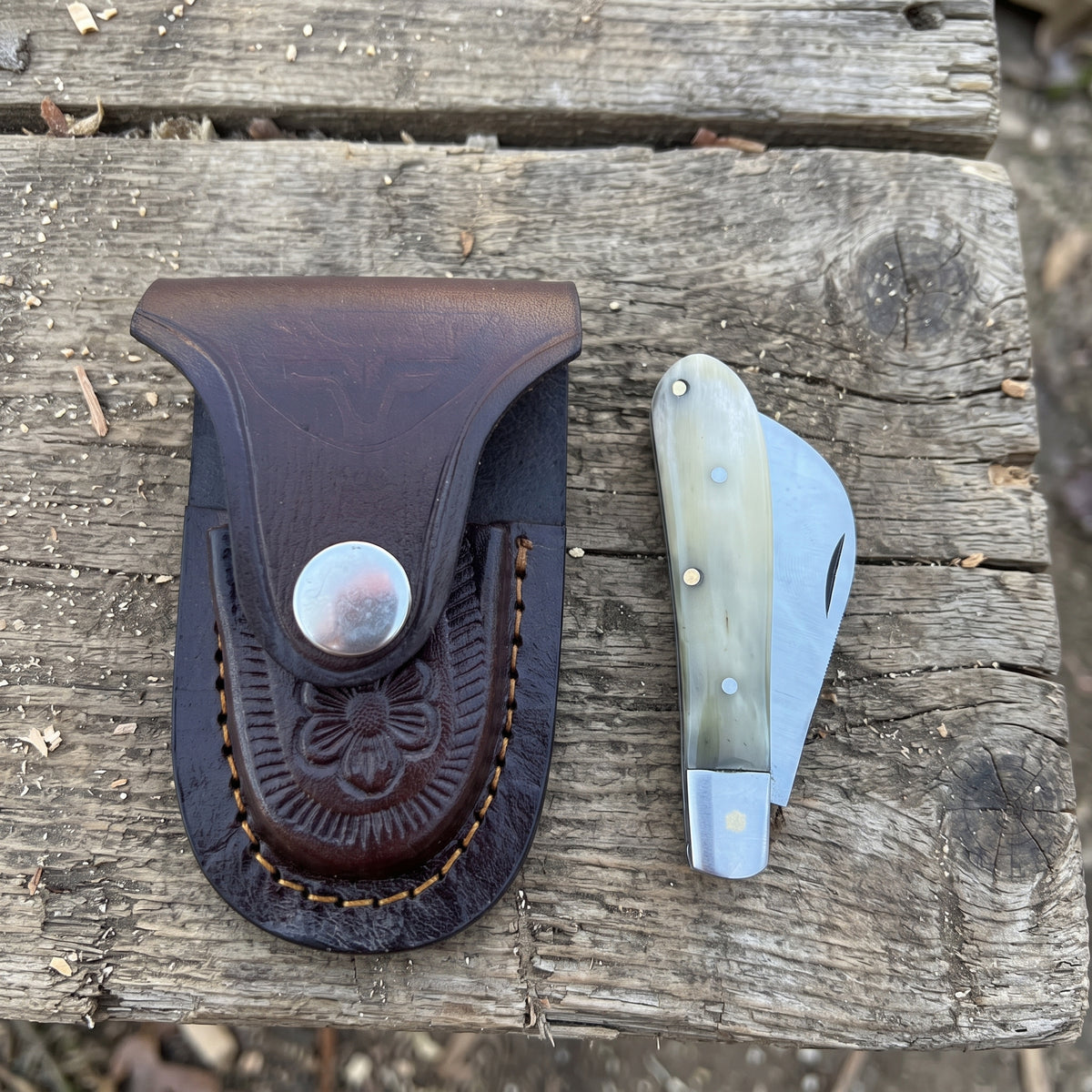 A dark brown leather folding knife sheath with an embossed floral pattern and snap closure, shown next to a folding pocket knife with a bone-colored handle on a weathered wooden surface.