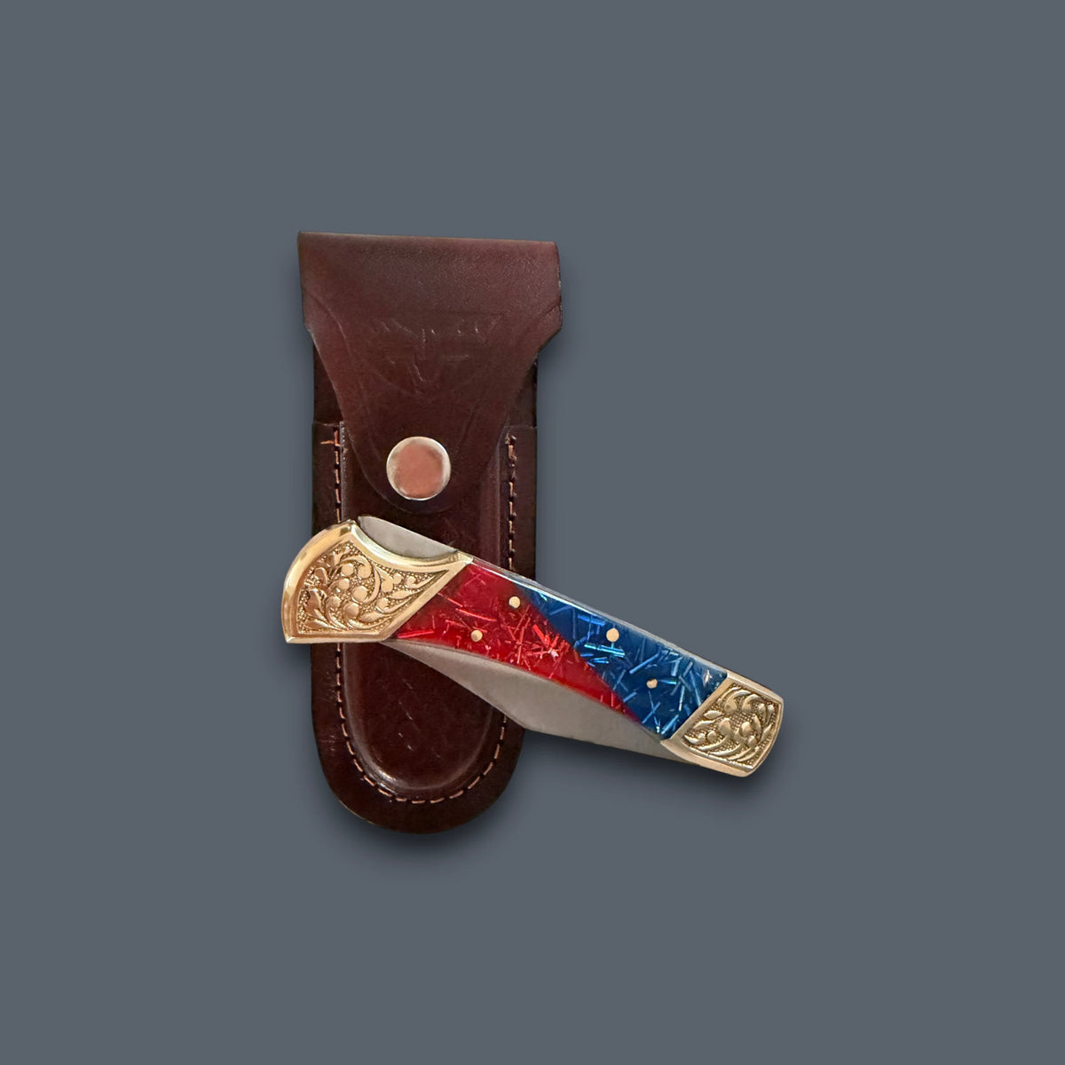A folding pocket knife with a red and blue inlay handle and gold-toned metal accents, shown partially inserted into a brown leather sheath against a dark gray background.