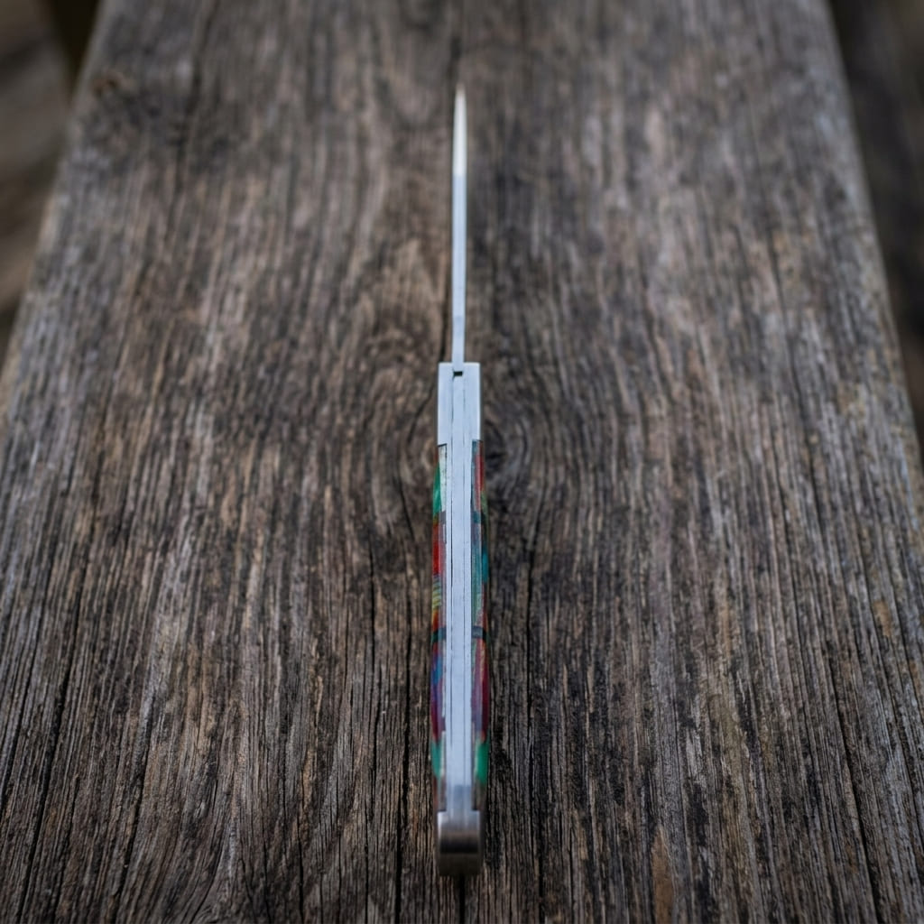 A old southern stainless steel pocket knife with a silver blade and a colorful inlay handle, standing vertically on a rustic dark wood surface.