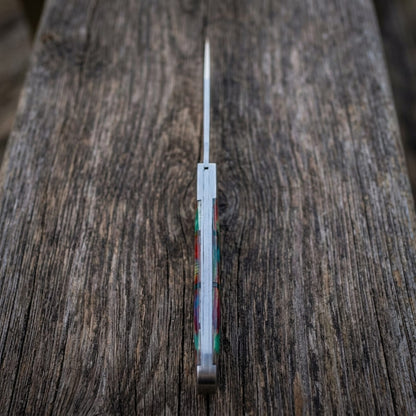 A old southern stainless steel pocket knife with a silver blade and a colorful inlay handle, standing vertically on a rustic dark wood surface.