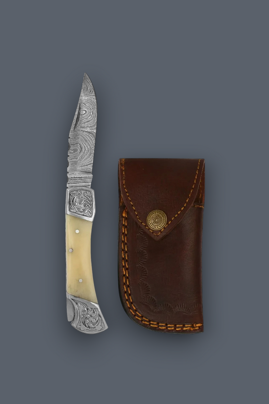 A decorative compact folding pocket knife with sheath with a patterned steel blade, cream-colored bone handles, and engraved silver metal fittings, shown next to its brown leather sheath on a solid dark gray background.