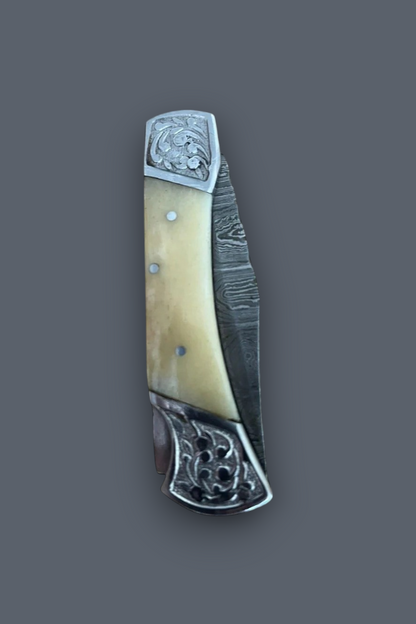 A folding pocket knife with a Damascus steel blade partially extended, featuring a cream-colored bone handle scale and ornate silver-toned metal fittings, set against a solid dark gray background.
