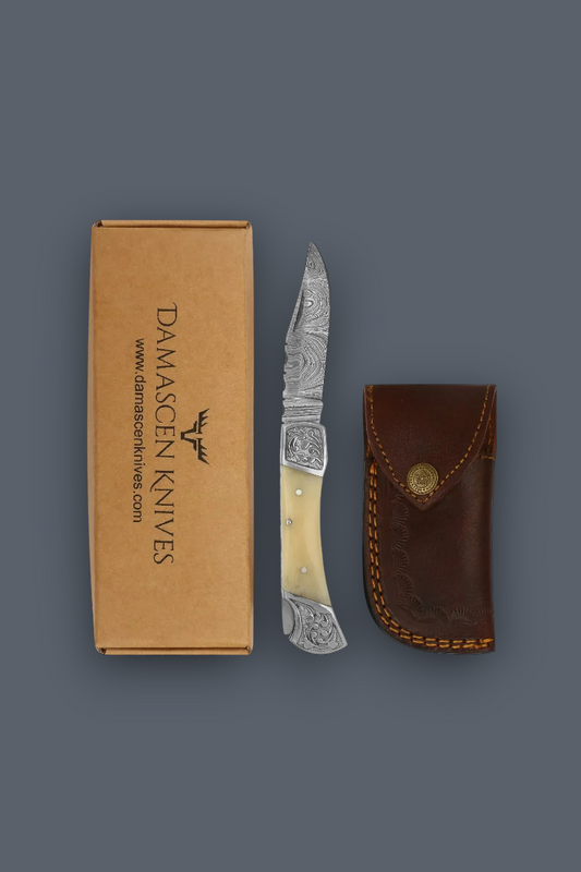 Damascen Knives folding pocket knife with an engraved silver blade and cream handle, shown next to its brown cardboard packaging box and a brown leather sheath on a dark gray background.