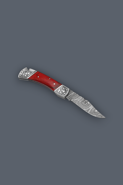 A folding pocket knife with a red handle and a patterned steel blade, shown diagonally on a solid dark gray background.